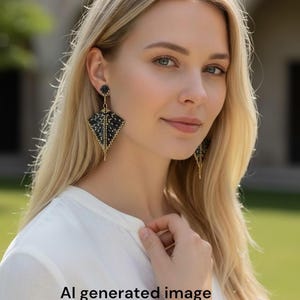 May include: Black and gold beaded earrings with a geometric kite shape. The earrings have a small round stud at the top and a long, thin gold chain at the bottom. The earrings are worn by a person with blonde hair.