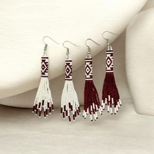 May include: Four beaded earrings with silver hooks. Two are white with burgundy accents, and two are burgundy with white accents. Each earring features a geometric pattern and a fringe of beads.