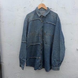 May include: A long-sleeve denim shirt in a medium blue wash featuring a button-down front, a classic collar, and two chest pockets. The shirt is constructed with horizontal panels of denim fabric that have visible, frayed raw-edge seams.