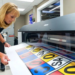 Op de afbeelding: Een vrouw bedient een grootformaatprinter. De printer print een vel papier met kleurrijke borden waarop nummers en het woord "SAS" staan.