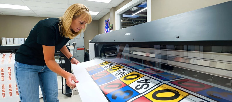 Op de afbeelding: Een vrouw bedient een grootformaatprinter. De printer print een vel papier met kleurrijke borden waarop nummers en het woord "SAS" staan.