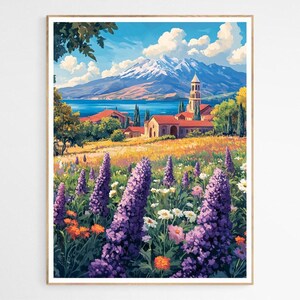May include: A framed art print of a colourful landscape. The artwork shows a field of purple and white wildflowers, a church, and a snow-capped mountain. The sky is blue with white clouds. The print is in a wooden frame.