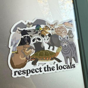 Peut inclure: Un autocollant représentant diverses illustrations d'animaux nord-américains, dont un raton laveur, un cerf, une tortue et un coyote. Le texte "respect the locals" est écrit en blanc et en gras.