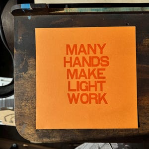 May include: Orange cardstock with the text "MANY HANDS MAKE LIGHT WORK" printed in bold, orange letters.