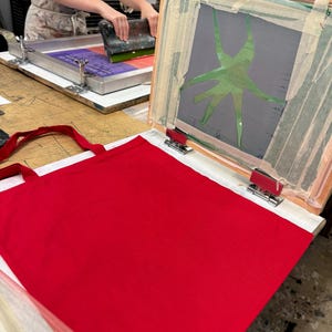 Pu&ograve; includere: Una borsa tote rossa &egrave; in fase di preparazione per la serigrafia. Il telaio per la serigrafia &egrave; aperto e mostra un design verde su uno sfondo grigio.