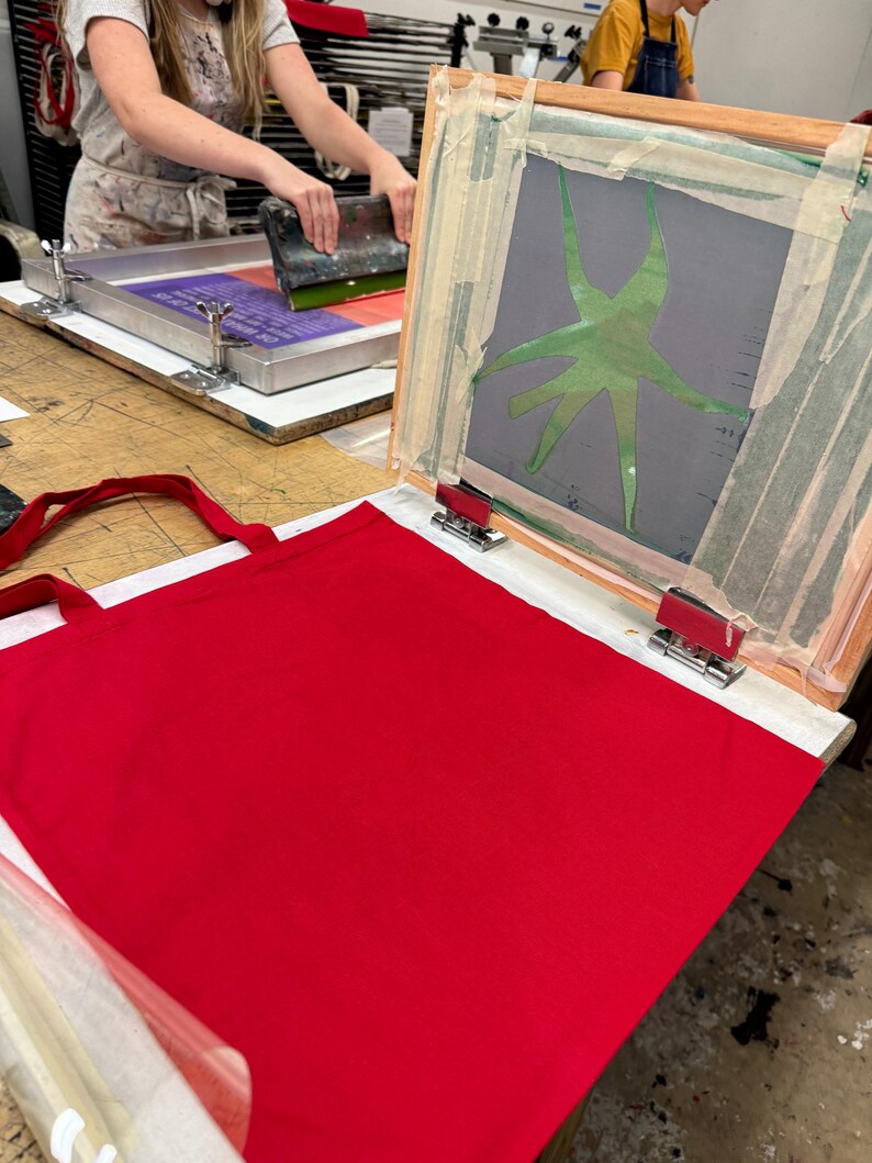 Pu&ograve; includere: Una borsa tote rossa &egrave; in fase di preparazione per la serigrafia. Il telaio per la serigrafia &egrave; aperto e mostra un design verde su uno sfondo grigio.