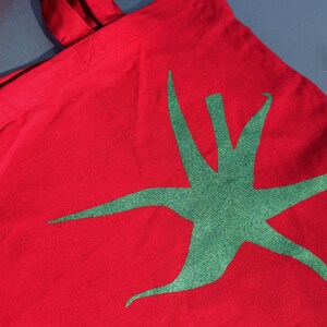 Pu&ograve; includere: Borsa tote in tela rossa con un design a stella verde.