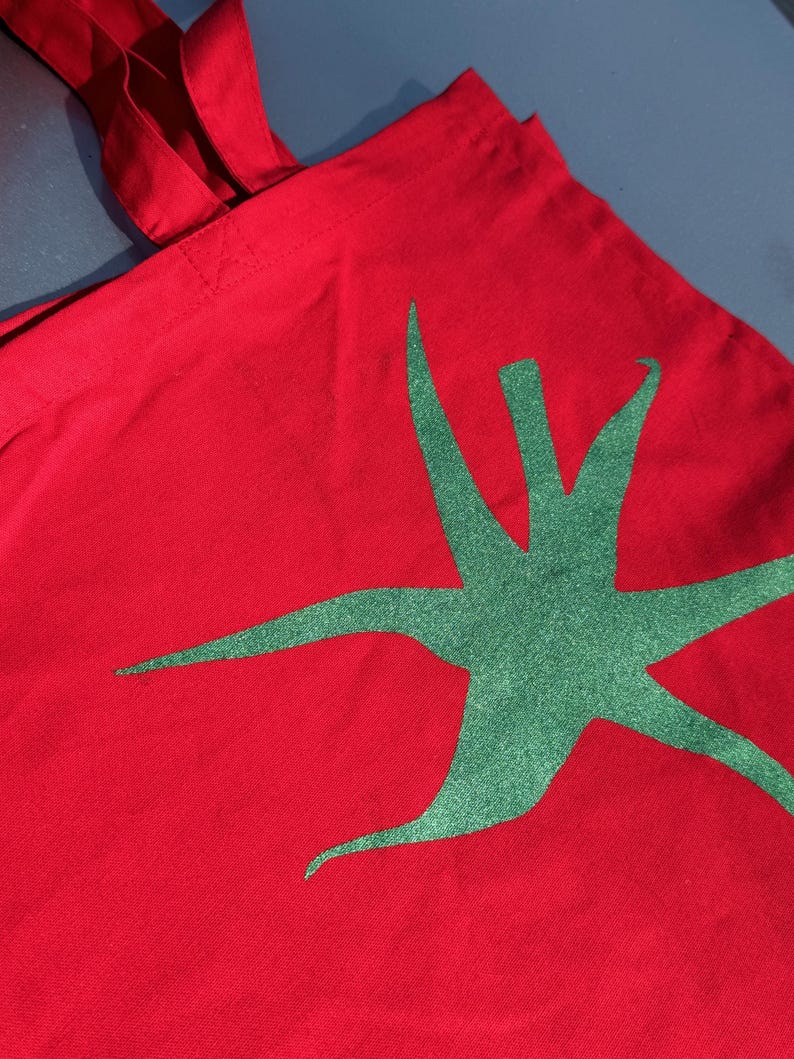 Pu&ograve; includere: Borsa tote in tela rossa con un design a stella verde.