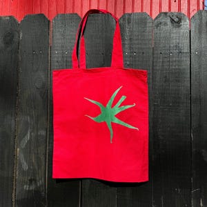 Pu&ograve; includere: Borsa tote in tela rossa con un design a stella stilizzato verde.