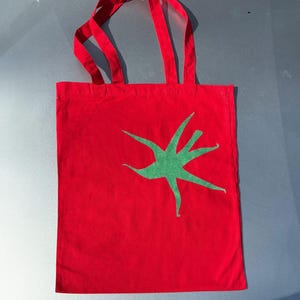 Pu&ograve; includere: Borsa tote in tela rossa con un design a stella verde.