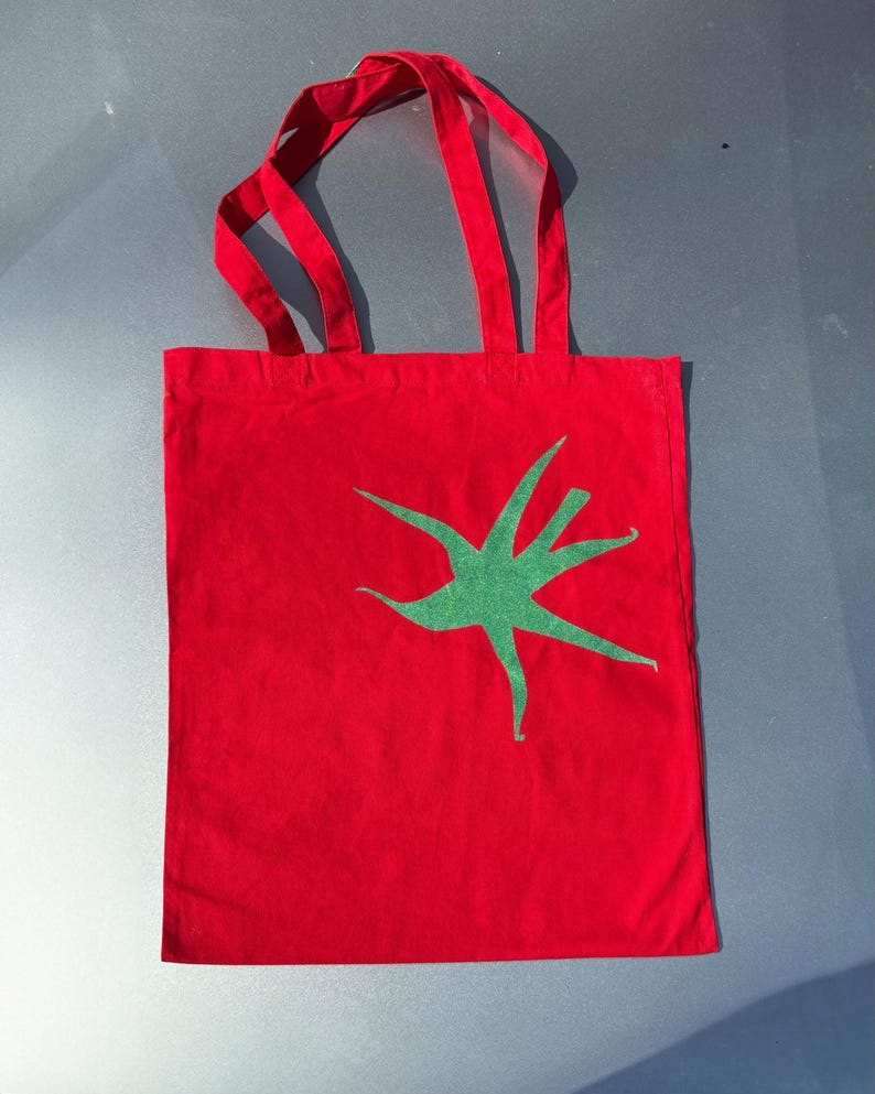 Pu&ograve; includere: Borsa tote in tela rossa con un design a stella verde.