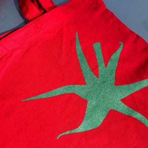 Pu&ograve; includere: Borsa tote in tela rossa con un design a stella verde.