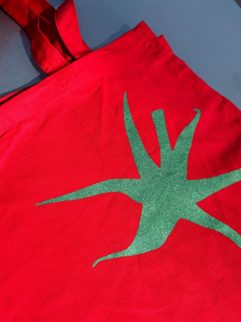 Pu&ograve; includere: Borsa tote in tela rossa con un design a stella verde.