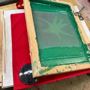 Pu&ograve; includere: Un telaio per serigrafia in legno con un disegno verde su un tessuto rosso. Il telaio ha la scritta "Speedball" stampata su di esso.