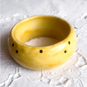 May include: A thick, rounded bangle bracelet with a glossy yellow glaze finish. The surface of the bracelet is decorated with small, evenly spaced black polka dots that wrap around the exterior circumference of the circular jewelry piece.