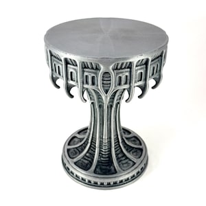 May include: A silver-colored, decorative pedestal table with an ornate, architectural design. The round tabletop is supported by a detailed, stylized base. The table is approximately 4 inches in diameter and 5 inches tall.