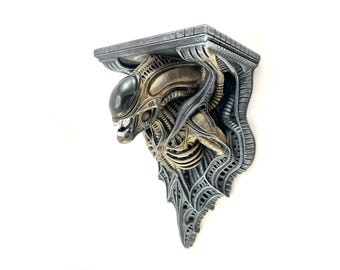 May include: A detailed, three-dimensional shelf featuring an Alien design. The shelf is primarily grey with bronze and black accents, showcasing the creature's head and body. The shelf is mounted on a white wall.