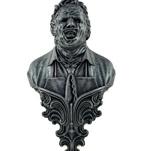 Pode incluir: Busto cinza escuro de um homem com um sorriso sinistro, usando uma camisa de colarinho e gravata, com uma base ornamental e decorativa. A escultura tem uma aparência texturizada e está sobre um fundo branco.