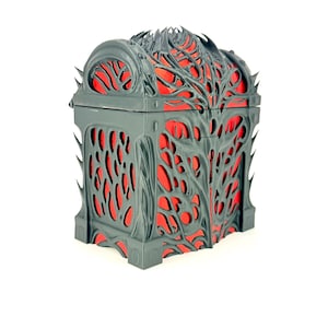 Op de afbeelding: Een decoratieve kist met een donkergrijze buitenkant en een felrode binnenkant. De kist heeft een ingewikkeld, organisch ontwerp met een roosterachtig patroon aan de zijkanten en bovenkant. De kist heeft een scharnierend deksel en een rechthoekige vorm.