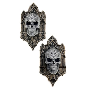 Pode incluir: Duas placas de parede em estilo gótico com design de caveira. As caveiras são cinzentas com órbitas escuras e estão sobre um fundo ornamental de cor bronze. As placas têm uma aparência detalhada e texturizada.