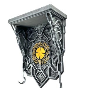 Puede incluir: Una estantería de pared gris con intrincados diseños geométricos y un elemento central dorado. La estantería tiene dos ganchos y detalles en forma de cadena, creando una solución de almacenamiento decorativa única.