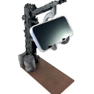 以下が含まれることがあります： 黒い3Dプリント製のスマートフォンスタンド。使い古された終末的なクレーンの構造と茶色の長方形のベースが特徴です。透明なケースに入ったスマートフォンを、暗い塔から伸びる機械的なアームで支えています。