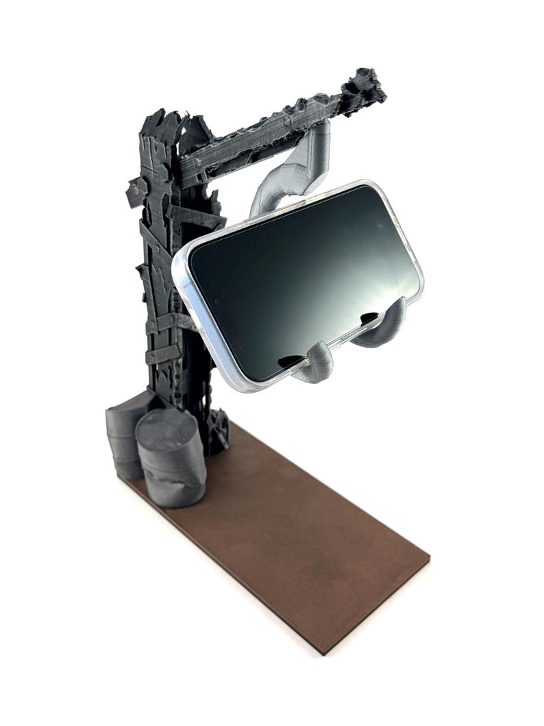 以下が含まれることがあります： 黒い3Dプリント製のスマートフォンスタンド。使い古された終末的なクレーンの構造と茶色の長方形のベースが特徴です。透明なケースに入ったスマートフォンを、暗い塔から伸びる機械的なアームで支えています。