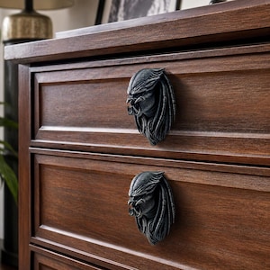 Puede incluir: Primer plano de una cómoda de madera marrón oscuro con dos cajones. Cada cajón presenta un tirador decorativo metálico con forma de rostro de Depredador. Los tiradores están detallados con líneas intrincadas y un acabado oscuro y envejecido.