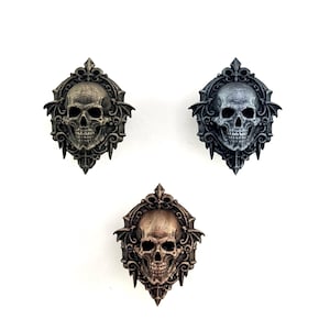 以下が含まれることがあります： ブロンズ、シルバー、ダークブロンズの3つの装飾的なスカルプラーク。各スカルは、コウモリのような翼と装飾的なボーダーを含む、精巧なディテールで縁取られています。スカルは白い背景に配置されています。