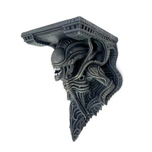 May include: A gray, sculpted shelf featuring the head and upper body of an Alien creature. The shelf has a rectangular top and detailed, textured design. The Alien's mouth is open, revealing teeth.