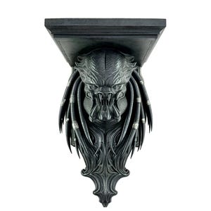 Puede incluir: Estantería de pared negra con temática Predator. La estantería presenta una cabeza de Predator esculpida en detalle con rastas y una expresión amenazante. La estantería tiene una parte superior rectangular y una base decorativa.