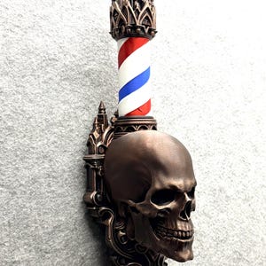 Puede incluir: Un poste de barbero montado en la pared con una base de cráneo humano de color bronce y detalles arquitectónicos góticos. El cilindro sobre el cráneo muestra el tradicional patrón de rayas diagonales en colores rojo, blanco y azul.