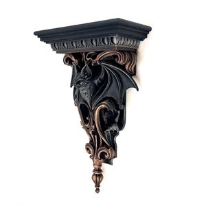 May include: A wall-mounted shelf featuring a flat top surface and an ornate bracket base. The bracket is sculpted with a detailed bat figure with spread wings, finished in a matte black color with metallic bronze highlights on the raised edges.