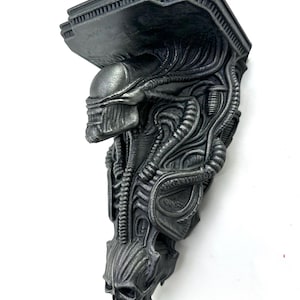 May include: A dark gray, wall-mounted shelf featuring a detailed, sculpted design of an Alien creature. The shelf has a rectangular top and a complex, textured body with a skull at the bottom. The shelf is made of a solid material.