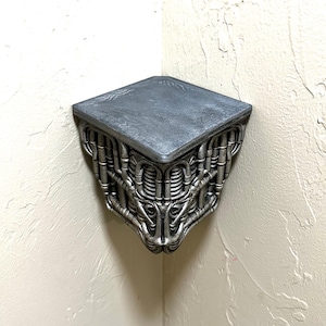 Puede incluir: Estantería de esquina plateada con un diseño industrial único. La estantería tiene una parte superior cuadrada y una estructura compleja en forma de tuberías que forma el soporte. El diseño recuerda al arte biomecánico.