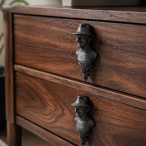 Op de afbeelding: Een donkerbruine houten dressoir met twee lades. Elke lade heeft een zwart, gebeeldhouwd handvat in de vorm van een man met een hoed. De houtnerf is zichtbaar, tegen een vage achtergrond.
