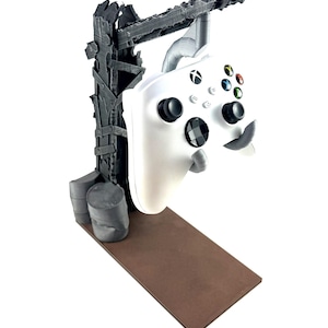 以下が含まれることがあります： 白のXboxワイヤレスコントローラーが、ダークグレーの荒廃したポスト・アポカリプス風スタンドに吊り下げられています。スタンドにはダメージ加工が施された垂直の梁、土台部分の2つの円筒形ドラム缶、そして平らな長方形の茶色のプラットフォームが備わっています。