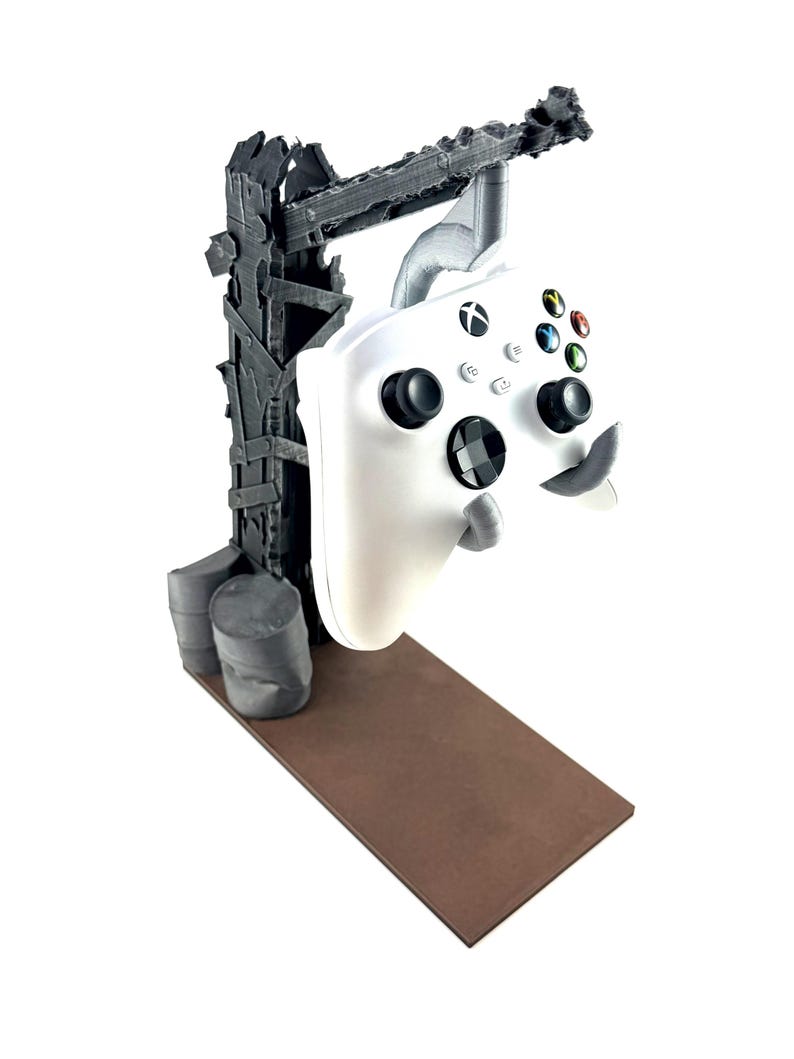以下が含まれることがあります： 白のXboxワイヤレスコントローラーが、ダークグレーの荒廃したポスト・アポカリプス風スタンドに吊り下げられています。スタンドにはダメージ加工が施された垂直の梁、土台部分の2つの円筒形ドラム缶、そして平らな長方形の茶色のプラットフォームが備わっています。