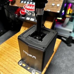 May include: A black Moza Racing gear shifter with a vertical handle and a rectangular base featuring a gated shift pattern. The unit is mounted on a wooden surface, with a Peterbilt branded control panel visible in the background.