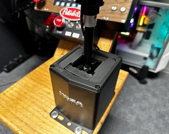 May include: A black Moza Racing gear shifter with a vertical handle and a rectangular base featuring a gated shift pattern. The unit is mounted on a wooden surface, with a Peterbilt branded control panel visible in the background.