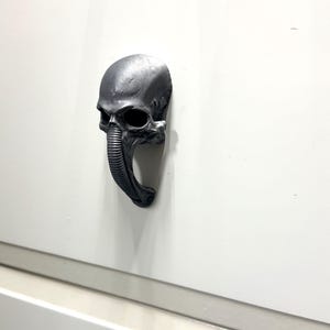 Pode incluir: Um puxador de armário em forma de caveira cinza metálico com um design alongado único. O puxador apresenta uma superfície texturizada detalhada e é montado em uma porta de armário branca. Este hardware decorativo adiciona um toque gótico ou industrial.