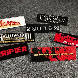 May include: A collection of seven horror movie themed iron-on patches. The patches are black with red and white lettering. The patches feature the titles "The Toxic Avenger", "The Texas Chainsaw Massacre", "Scream", "Halloween III: Season of the Witch", "Terrifier", "A Nightmare on Elm Street", and "The Texas Chain Saw Massacre".