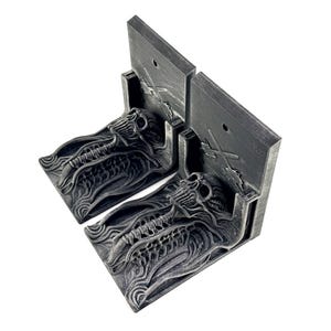 May include: A pair of grey bookends featuring a skull and skeletal design. The bookends have a detailed, textured appearance and a rectangular back with a small hole and an etched airplane design.