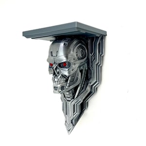 May include: A grey metal wall shelf in the shape of a Terminator skull with red eyes. The shelf has a rectangular top and a detailed skull base, ideal for displaying items or adding a unique touch to a room. The shelf is made of metal.