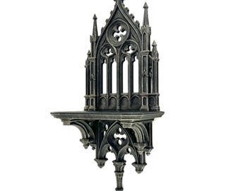Puede incluir: Una estantería de pared de estilo gótico en gris oscuro con intrincados detalles arquitectónicos. La estantería presenta ventanas arqueadas, pináculos puntiagudos y tallas decorativas, evocando una estética de catedral medieval. Tiene una pequeña superficie plana.