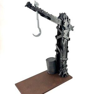 以下が含まれることがあります： 突き出したアームからフックが吊り下げられた、ダークグレーの風化したクレーン構造物。土台は平らな長方形の茶色のプラットフォームで、クレーンの足元には小さな円筒形のダークグレーの樽が配置されています。