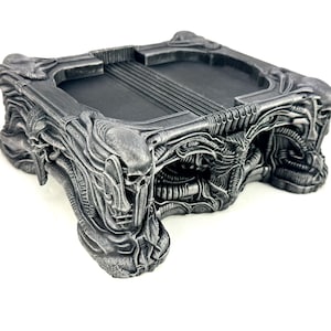 May include: A detailed, gray, rectangular tray with an intricate, biomechanical design. The tray features a central recessed area with ridged detailing and is supported by sculpted, organic-looking legs. The overall aesthetic is reminiscent of science fiction.