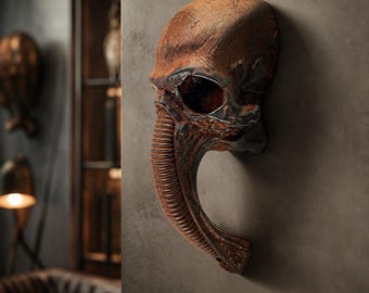 May include: A detailed, brown wall-mounted sculpture of an alien skull with a long, ribbed proboscis. The sculpture has a weathered, textured appearance, with a large eye socket and a dark interior. The overall aesthetic is reminiscent of science fiction.