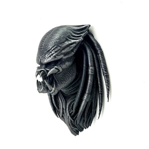 Pode incluir: Escultura detalhada de máscara Predator cinza metálico. A máscara apresenta texturas intrincadas, dentes afiados e dreadlocks, montada em um fundo branco. A escultura é uma representação detalhada do icônico personagem do filme.