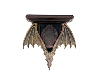 Può includere: Una mensola da parete color bronzo con un design gotico. La mensola presenta una parte superiore marrone scuro, un pannello centrale ad arco e ali di pipistrello stilizzate che si estendono verso l'esterno. La parte inferiore della mensola ha una creatura simile a un pipistrello.